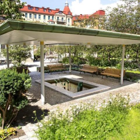 A roof with an opening in the middle over a small pond and a few benches, surronded by bushes, trees and lawns.